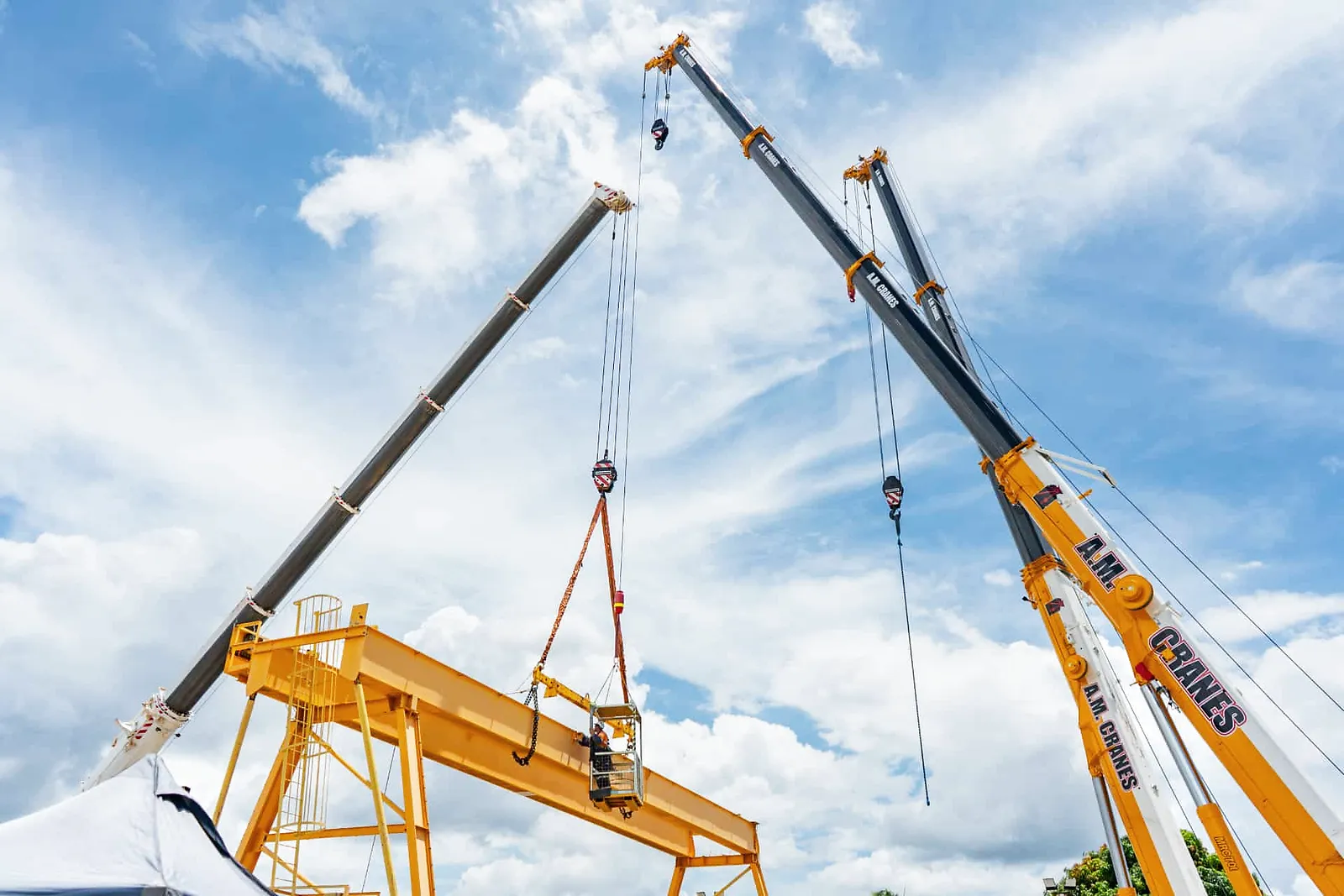 Two mobile cranes lifting a yellow gantry crane beam into place under a blue sky