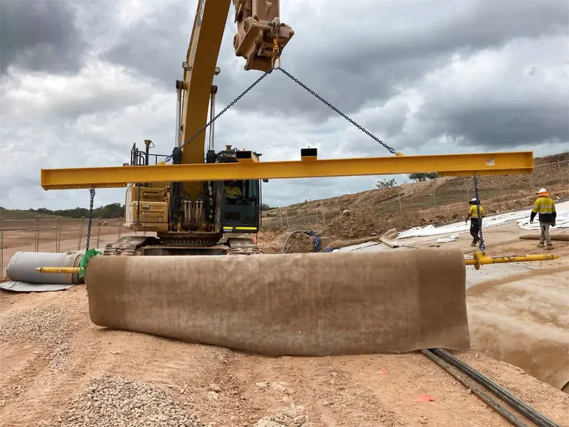 A yellow spreader beam lifting a large load on a red dirt work site